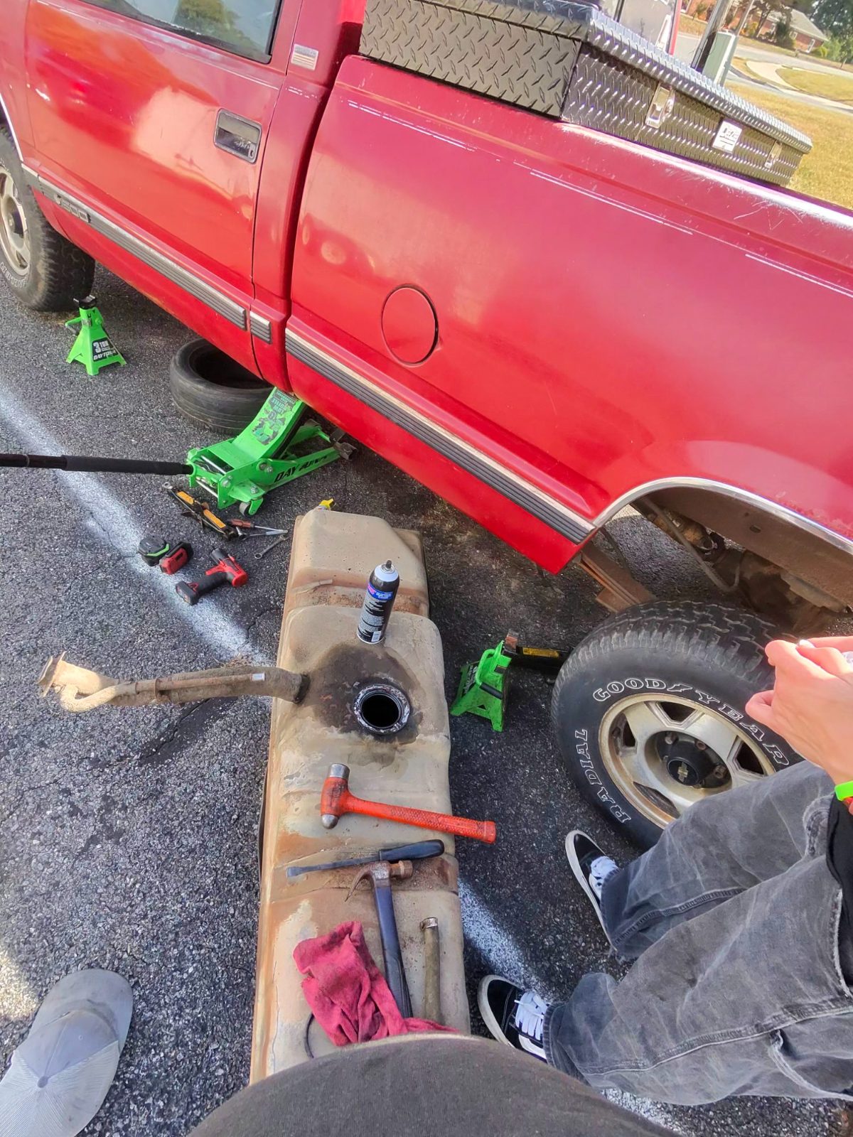 Fuel tank dropped from a red pickup on jack stands with tools spread on the pavement